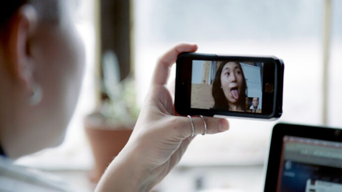 A young woman's hand holding a phone to record herself on video; the screen shows her sticking her tongue out in a playful face.